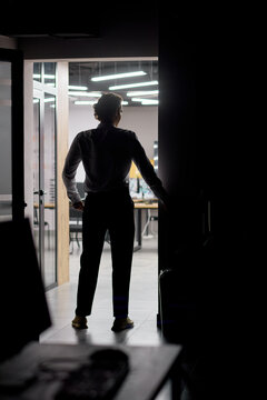 Silhouetted man opening glass door into brightly lit modern office interior at night, entering contemporary workplace with desks, chairs, overhead lighting and strong backlit contrast