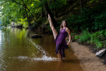 Fototapeta premium Young Woman Dancing on River Shore in Purple Dress Showing High Kick and Splash in Nature