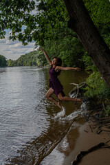 Fototapeta premium Young Woman Jumping Into River From Tree-Lined Shore While Splashing Water In Summer