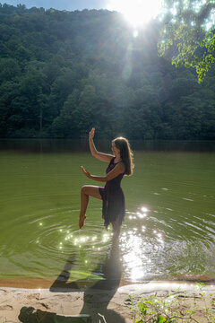 Woman Dancing In Shallow Lake At Sunset With Sunlight Reflection And Forest Background