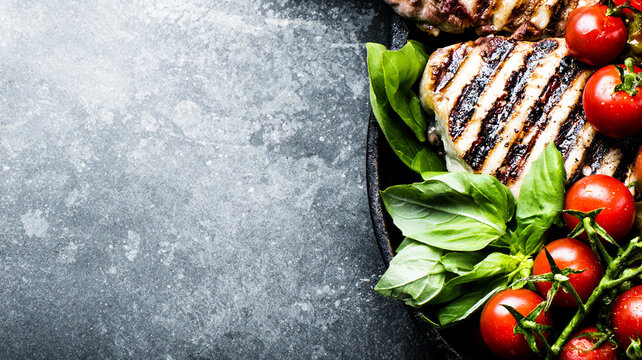 Grilled meat steaks with fresh cherry tomatoes and basil leaves on a dark stone background