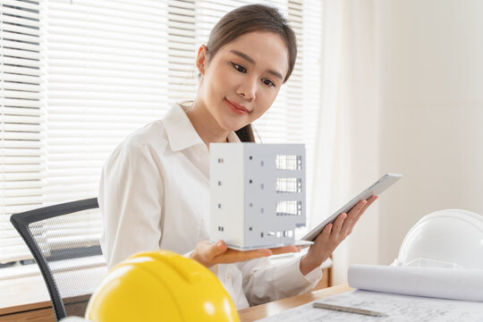 Professional young Asian woman engineer inspecting house model with tablet and construction safety helmet on workspace desk