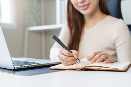 Happy Asian woman working on laptop computer and writing in notebook in modern home office concept for remote work and online learning