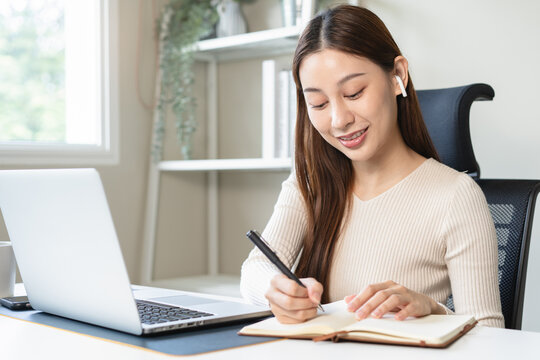 Happy Asian woman working on laptop computer and writing in notebook in modern home office concept for remote work and online learning