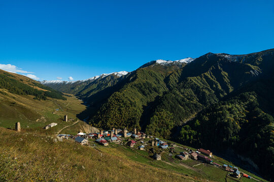 High angle landscape photograph of remote Adishi village with traditional Svan towers nestled in mountain valley under clear blue sky in Svaneti Georgia.