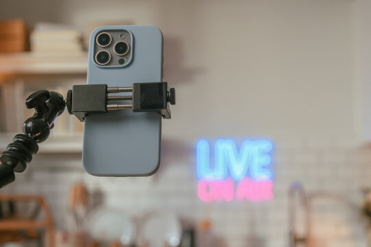 Closeup of a smartphone clamped on a flexible tripod arm for live streaming, with a glowing Live On Air neon sign blurred softly in a warm indoor background creator gear in focus.