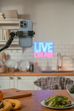 Closeup of a smartphone clamped on a flexible tripod arm for live streaming, with a glowing Live On Air neon sign blurred softly in a warm indoor background creator gear in focus.