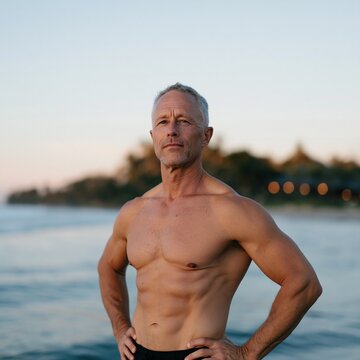 Fit shirtless man standing by ocean at dusk with hands on hips for fitness background and wellness banner design