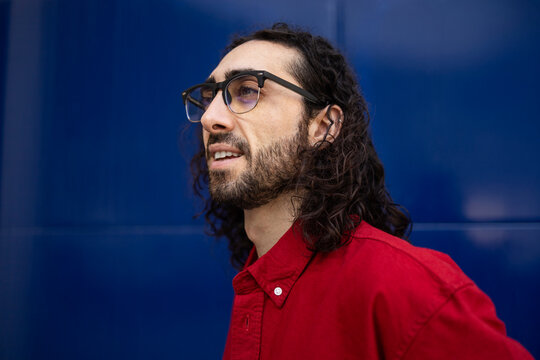 Bearded man with glasses in red shirt against blue wall