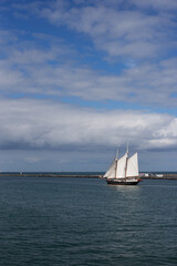 Сlassic black two-masted wooden schooner with white sails navigating sea water near stone pier breakwater under dramatic cloudy sky. © Olena