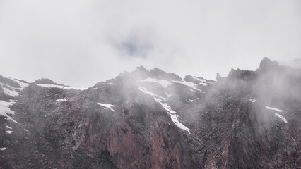 Jagged granite cliffs and lingering snowfields emerge through drifting mountain fog above Bogdanovich Glacier in the high alpine landscape of Kazakhstan. © Kolchanov