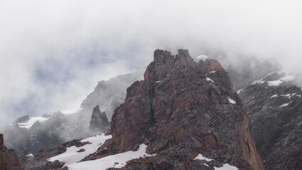 Jagged granite cliffs and lingering snowfields emerge through drifting mountain fog above Bogdanovich Glacier in the high alpine landscape of Kazakhstan. © Kolchanov