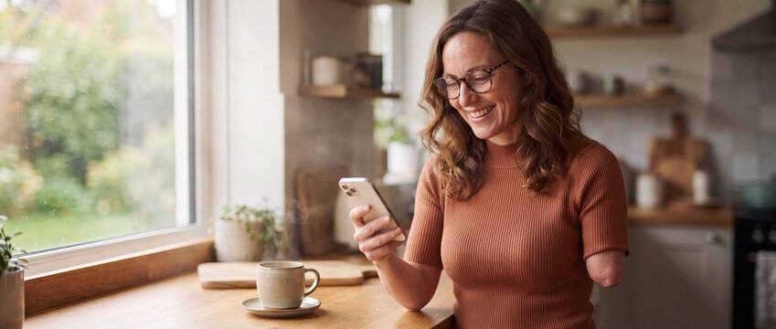 Amputee woman using smartphone in kitchen while smiling and enjoying hot coffee at a bright wooden counter by the window, conveying inclusion, domestic lifestyle, and mobile communication