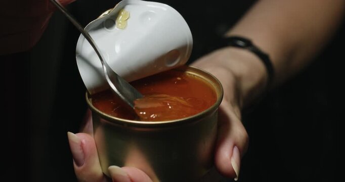 A close-up of a woman using a teaspoon to take a portion of jelly and pate to feed her pet cat.