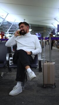Elegant man sitting in an airport departure lounge with his luggage, talking on his smartphone. Vertical video with copy space.