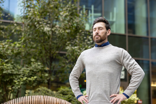 Young businessman standing near office center outside, holding hands on waist, eyes closed and resting