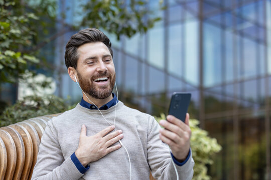 A smiling young man wearing headphones is sitting on a bench outside and having an online conversation on his phone camera