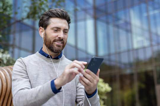 Close-up photo of a young man wearing headphones sitting on a bench outside and using a mobile phone