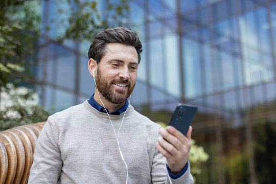 A young man sitting on the street wearing headphones looks into the camera of a mobile phone he is holding in his hand