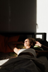Fototapeta premium Relaxing woman enjoying morning sunlight in a cozy bedroom, soaking in warmth and comfort while lying on a soft bed with pillows and blankets