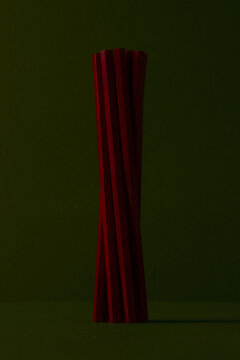 An elegant abstract composition featuring a twisted bundle of deep red hexagonal pencils standing vertically against a dark olive green background with subtle lighting and dramatic shadows.
