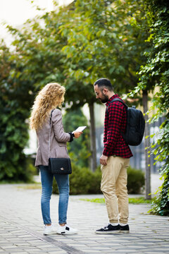 Young couple standing on a city sidewalk, engaged in conversation while facing each other, surrounded by greenery and trees. Casual fashion with jeans and backpacks highlighted in the urban setting.
