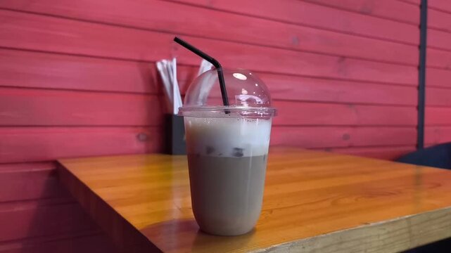 Close-up shot of a refreshing iced latte in a clear takeaway cup placed on a caf&eacute; table inside a stylish coffee shop with striking red walls. The bold red interior