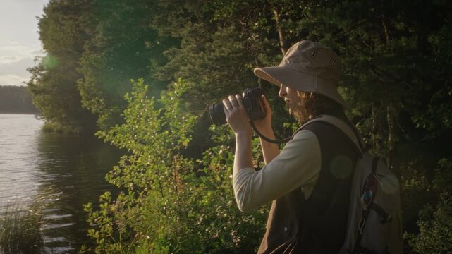Young adult Caucasian woman with backpack standing near lake, raising digital camera to look at forest landscape. Footage is ideal for travel, outdoor gear, and tourism marketing campaigns