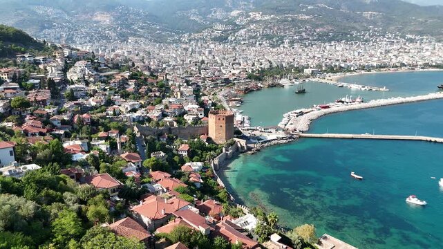 A breathtaking panoramic aerial video capturing the historic Red Tower (Kızıl Kule), the bustling harbor of Alanya, and the Mediterranean cityscape. Numerous tourist pirate ships, yachts, and motorboa