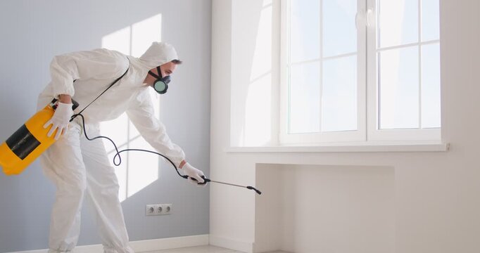 Pest control service works, exterminator in protective suit spraying cockroach insecticide for empty room disinfection. Worker with respirator mask holding sprayer to apply pesticide near window