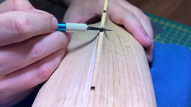 Hands carefully applying center planking line on wooden scale model boat hull, meticulous craftsmanship, DIY boat modeling.