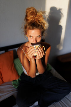 Woman enjoying a warm drink while sitting on her bed in a cozy atmosphere during the morning sunlight