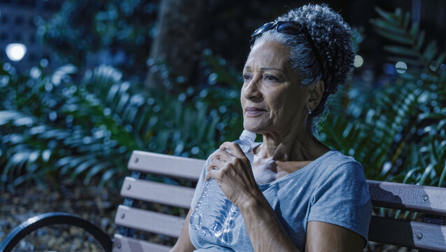 Senior woman sitting alone on park bench at night with a bottle of water