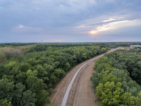 Cycling path curving along a flood dyke through green floodplain forest under a pastel sunset sky, aerial view