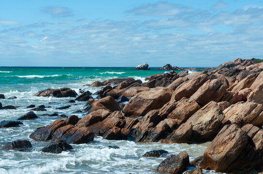 Rocky coastline with turquoise ocean waves, granite boulders and a wide blue sky with soft clouds