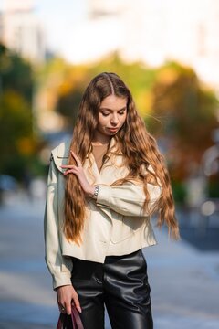 Young woman portrait on city street in autumn sunlight, wearing beige jacket and black trousers with long wavy hair, calm expression, soft bokeh and urban fashion mood