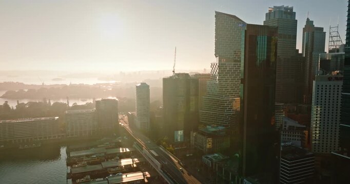 Sydney Central Business District buildings and skyscrapers rising over Circular Quay and harbor waters, illuminated by warm morning sunlight, urban environment and modern architecture. Aerial shot