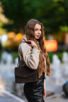 Young woman outdoors wearing beige coat brown shoulder bag fashion portrait park warm autumn light blurred background soft focus backdrop vibrant foliage casual chic styling urban street vibe