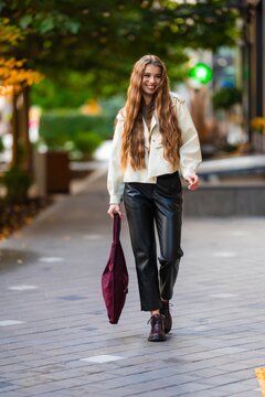 Young woman strolling along sunlit city sidewalk in beige jacket and black leather pants, showcasing urban chic style amid autumn foliage and lively downtown setting