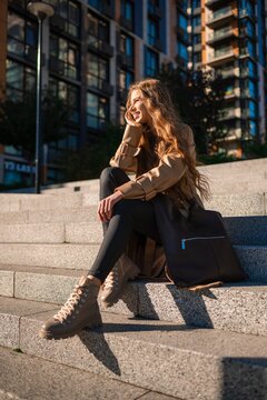 Young woman seated on city steps wearing beige trench coat, black boots, and carrying large tote bag beside modern glass buildings in sunlit urban scene with casual street style mood