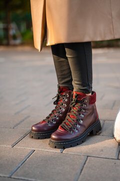 Beige coat hem layered above black leggings with burgundy leather hiking boots on sunlit city pavement, creating stylish autumn street fashion with rich texture and warm color