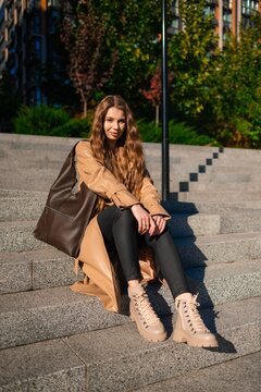 Young woman seated on granite steps in city park wearing beige trench coat and large brown backpack, posing in casual urban fashion under sunny daylight with greenery and modern architecture