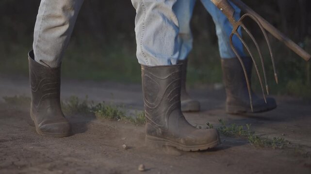 Farmer boots pitchforks road. A man walks along a dirt path, wearing boots while holding a pitchfork. Another man follows closely behind, both leaving dust in their wake.