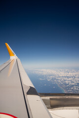 Airplane wing with yellow tip flies over coastline and sea under clear blue sky. High altitude flight view frames air travel tourism and commercial aviation concept. © Tetiana