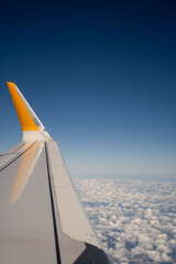 Airplane wing with yellow tip flies high above white cloud layer against blue sky. High altitude flight view frames air travel tourism and commercial aviation concept. © Tetiana