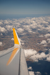 Yellow wingtip of passenger airplane flying high above white clouds and landscape during day. Air transportation and commercial flight route concept. © Tetiana