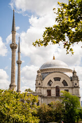 Historic mosque building with tall minarets and dome framed by green tree leaves under cloudy sky. Traditional Ottoman architecture and Turkish cultural heritage concept. © Tetiana