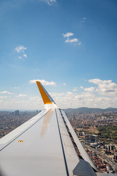 Airplane wing with yellow winglet flies high above the city landscape under blue sky. Commercial aviation travel and international flight transport concept.