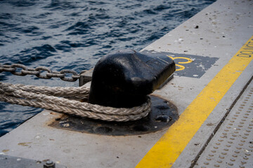 Close up view of heavy black mooring bollard with thick rope secured on dock near water. Marine transportation concept depicting port infrastructure safety and maritime travel background. © Tetiana