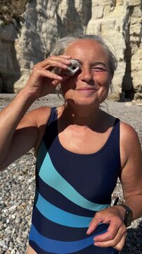 Mature smiling woman in swimwear looking through hag stone hole towards camera on pebble beach.